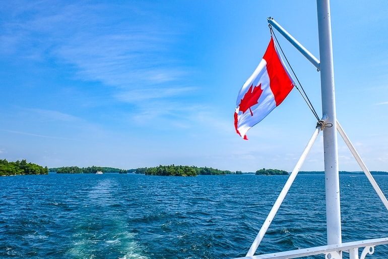 canada flag on white pole flying off back of boat with blue water below.