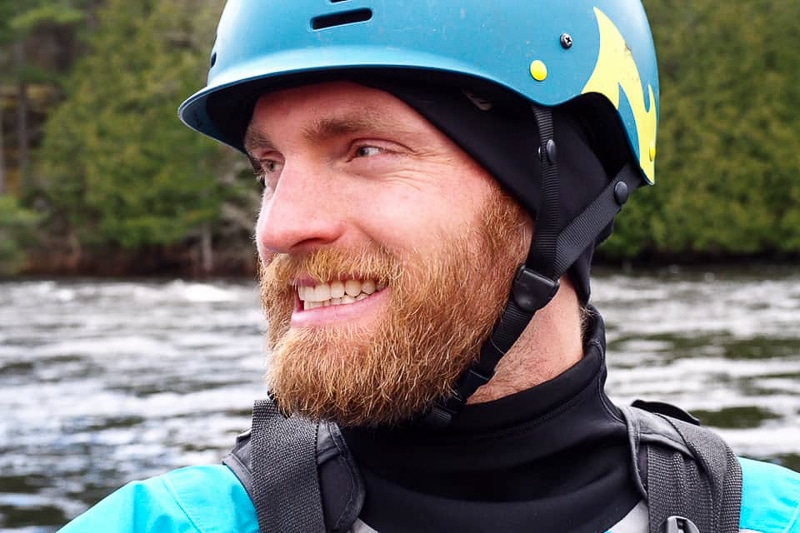 smiling man with helmet and life jacket on with river behind.
