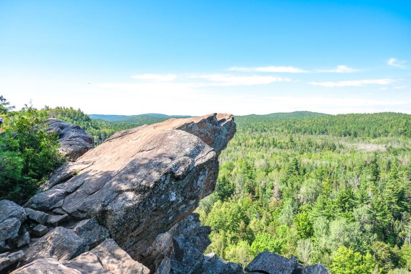 large rock extending off cliff over top of green trees in below valley.