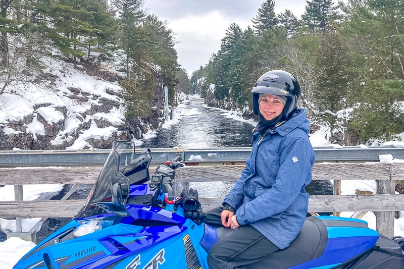 woman with helmet in blue coat sitting on snowmobile with river behind.